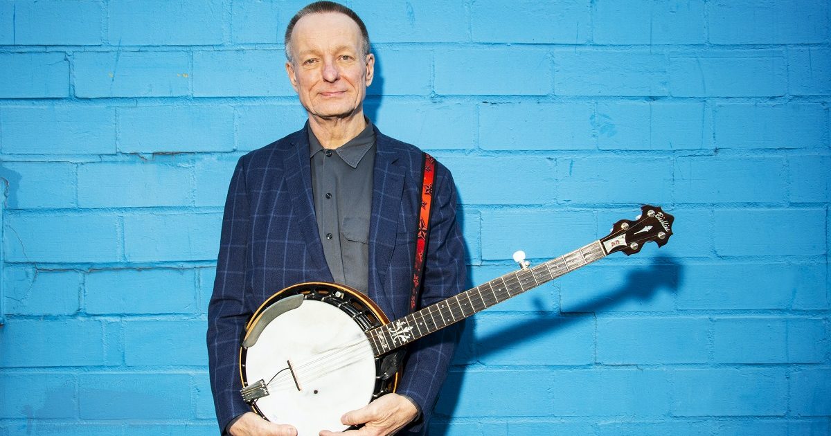 danny barnes standing in front of a blue wall with a banjo