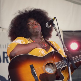 British singer/songwriter Yola's first Newport Folk Festival. Yola, wearing a yellow dress, places her hand over her heart while holding a guitar at Newport Folk Fest.
