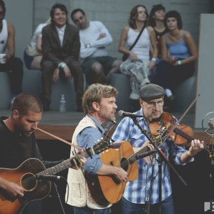 Willie Watson and band, featuring Gabe Witcher on fiddle, perform while other artists watch in the background.